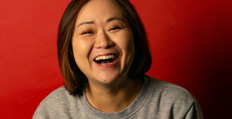 Comedian He Huang smiling against a red background. Photography by Teniola Komolafe.